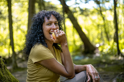 Smiling mature woman biting an apple and looking at the camera while sitting in forest.