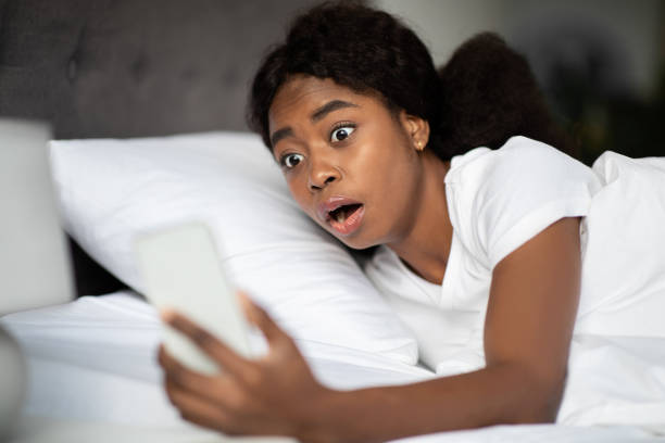 donna nera scioccata che guarda il cellulare, sdraiata a letto - dormire troppo foto e immagini stock