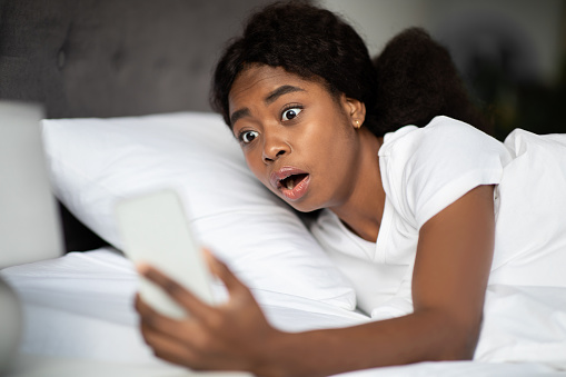 Shocked african american young woman laying in bed and looking at smartphone screen in her hand at home, overslept. Black lady is late for appointment or work or class study, copy space