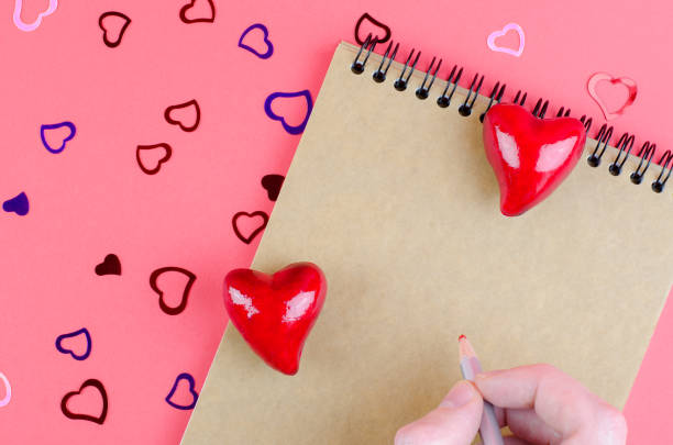 istock Part of a notepad on a pink background with hearts with space for text. Man's hand with a pencil on a notebook. Valentine's day concept 1368566276