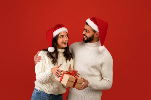 weihnachten, neujahrsgeschenke. aufgeregter arabischer typ, der seiner frau weihnachtsgeschenk macht, weihnachten zusammen feiert, weihnachtsmützen trägt - gift new year couple men stock-fotos und bilder