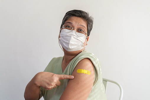 Portrait of senior Asian woman with eyeglasses sitting against white background, showing and pointing her arm with yellow bandage after got vaccinated, smiling and looking at camera.