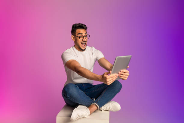 istock Emotional young Arab man sitting cross legged with digital tablet, looking at screen in shock, neon light 1368568485