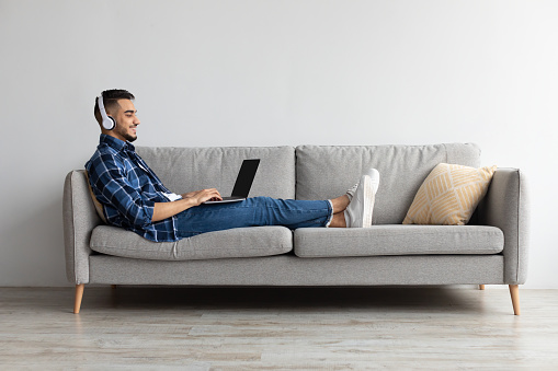 Happy Arab guy resting sitting on couch at home, watching movie on laptop, using pc in wireless headset. Cheerful man playing video games typing on keyboard relaxing on weekend in living room