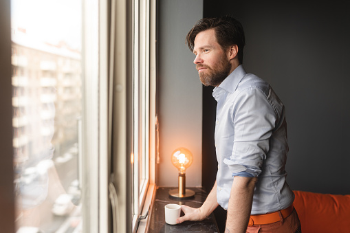 A man at home alone in his apartment. He is standing by the window, looking out on the city.