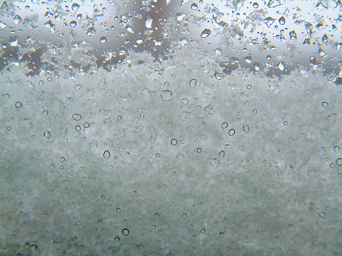 A macro shot of snow on a window sill, with water droplets visible on the glass. Both the snow build up and water droplets are in focus.Background through the glass is nicely blurred with DOF.