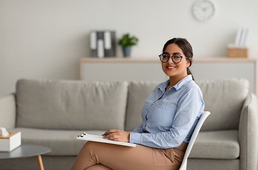 Psicóloga árabe segura de sí misma escribiendo en portapapeles, sentada en una oficina moderna y sonriendo ante la cámara, espacio libre photo