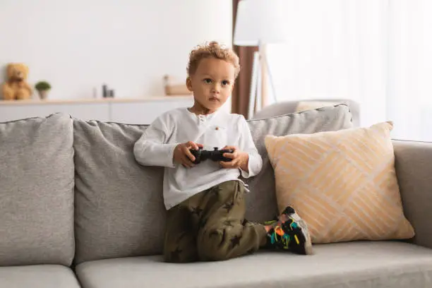 Black Little Boy Playing Videogames Using Gamepad Controller Having Fun Sitting On Sofa At Home On Weekend. Child's Leisure And Entertainment. Video Gaming Concept