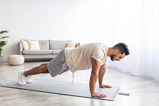Handsome young Arab man doing plank, following online video tutorial on smartphone at home, full length. Athletic middle Eastern guy exercising indoors during coronavirus lockdown