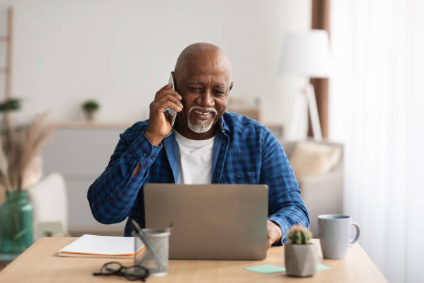 mature african man talking on cellphone using laptop in office - calling imagens e fotografias de stock