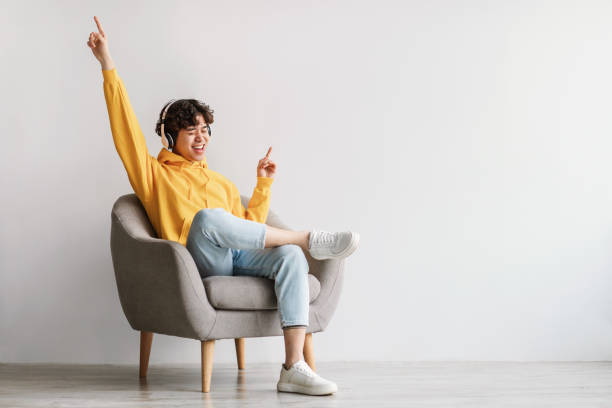 Joyful Asian guy in headphones sitting in armchair, listening to music, dancing to favorite song against white wall Joyful young Asian guy in wireless headphones sitting in armchair, having fun, listening to music, raising his arm, dancing to favorite song, enjoying cool soundtrack against white wall, empty space asian man headphones stock pictures, royalty-free photos & images