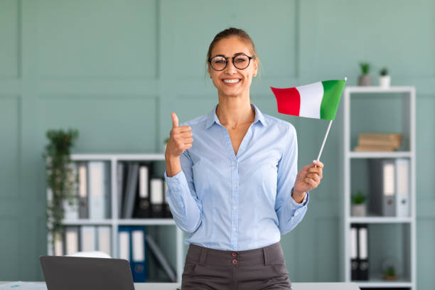 feliz tutora con la bandera de alemania mostrando el pulgar hacia arriba, de pie cerca del lugar de trabajo en la oficina y sonriendo a la cámara - italiano idioma fotografías e imágenes de stock