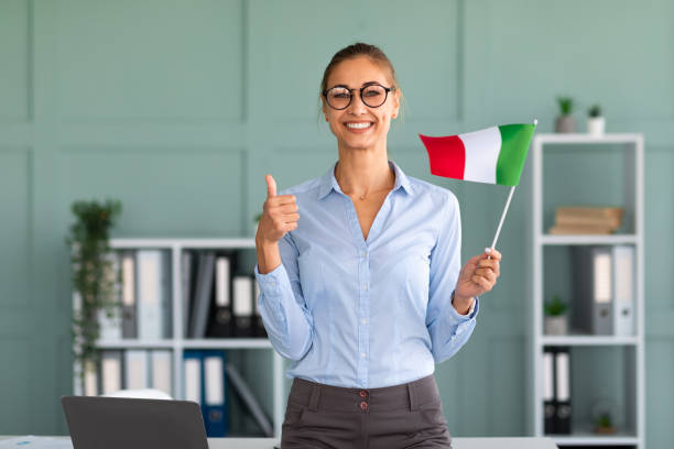 happy female tutor with flag of germany showing thumb up, standing near workplace in office and smiling at camera - 意大利語 個照片及圖片檔