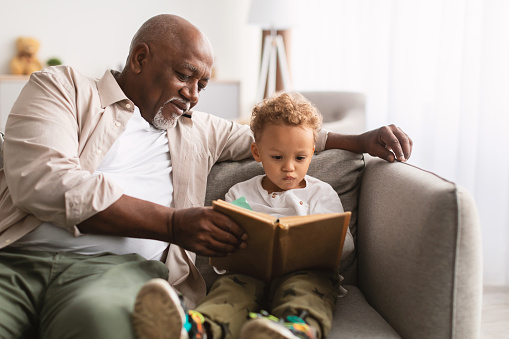 African American Grandfather And Little Grandson Reading A Book Sitting Together On Sofa At Home. Senior Male Teaching His Grandchild To Read On Weekend. Family Moments Concept