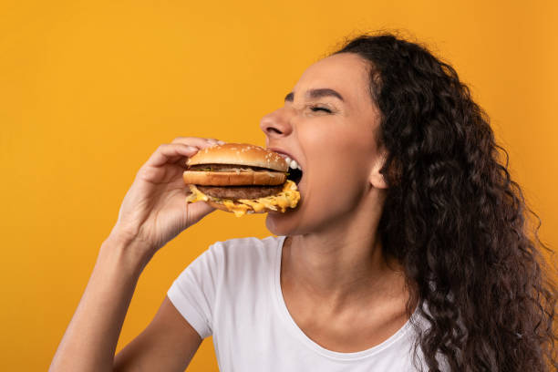 Funny Hungry Latin Lady Holding Burger Biting Sandwich At Studio Gluttony. Closeup Portrait Of Funny Hungry Lady Biting Burger Eating Junk Food Posing Over Yellow Orange Studio Background. Woman Enjoying Big Hamburger. Unhealthy Nutrition And Bad Eating Habit fast food restaurant photos stock pictures, royalty-free photos & images