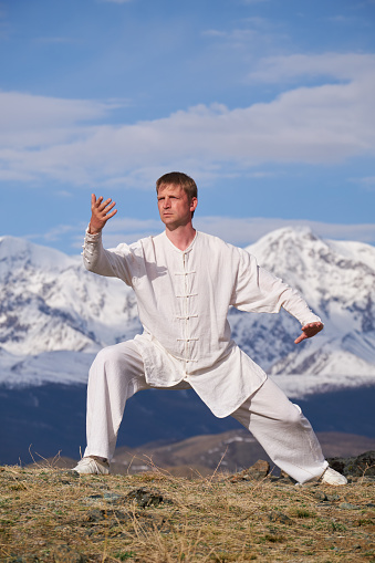 Wushu master in a white sports uniform training on the hill. Kungfu champion trains maritial arts in nature on background of snowy mountains.