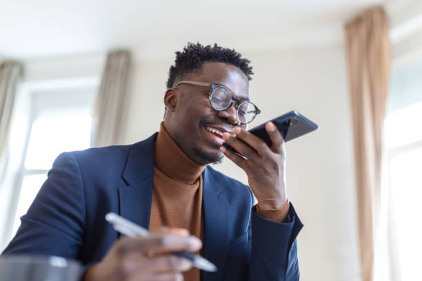 istock Arfican business man writing notes in notepad while talking by speakerphone from workspace. speech recognition concept. hands-free communication. 1368576760