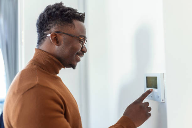 Smiling African American man using modern smart home system, controller on wall, positive young man switching temperature on thermostat or activating security alarm in apartment Smiling African American man using modern smart home system, controller on wall, positive young man switching temperature on thermostat or activating security alarm in apartment thermostat stock pictures, royalty-free photos & images