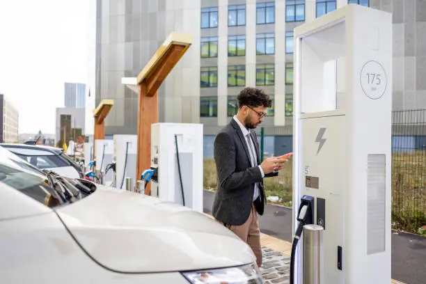 A wide-angle side view of a man in business dress using his mobile phone to access an app to be able to put his electric vehicle on charge at an electrical charging station in Newcastle City Centre in the North East of England. It is in a row of electric charging points.