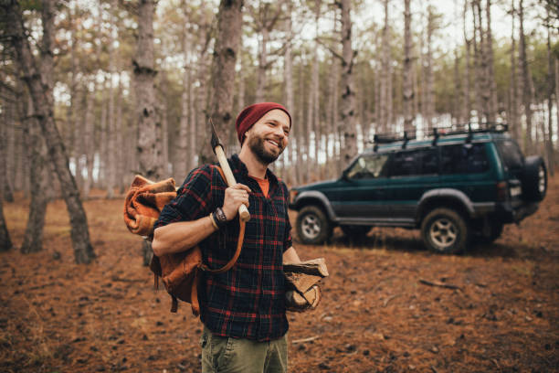 istock Millennial man hipster with beard wearing backpack, holding axe and firewood in forest with car on background. 1368580647