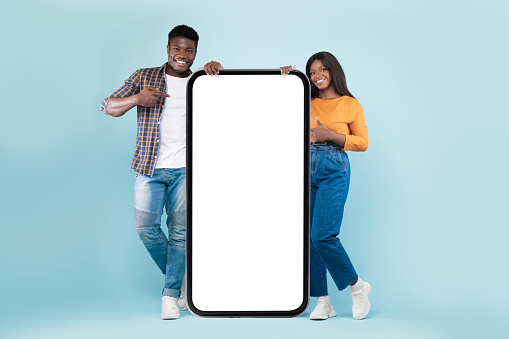 Great Mobile App. Excited black couple pointing at big giant smartphone with mock up copy space, promoting application or website, advertising product or service, blue studio wall. Full body length