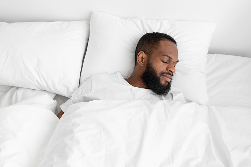 Calm millennial black bearded male sleeping on bed, soft pillow with white duvet in bedroom enjoy rest on weekend and vacation, empty space. Healthy sleep, health care and relax at home, top view