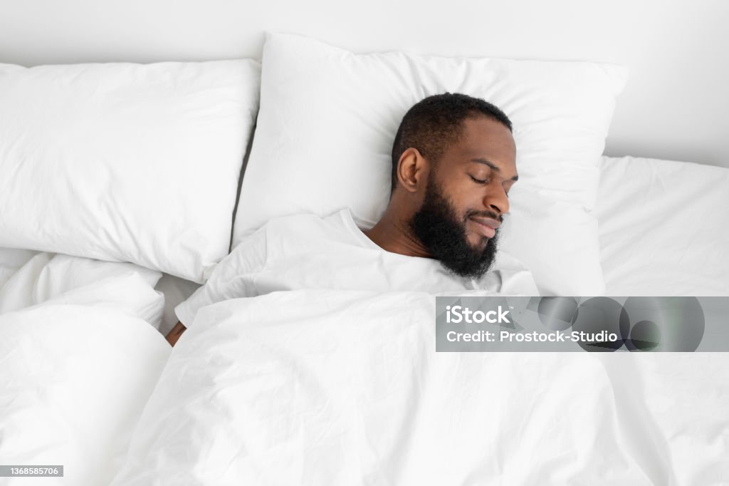 Calm millennial black bearded male sleeping on bed, soft pillow with white duvet in bedroom enjoy rest Calm millennial black bearded male sleeping on bed, soft pillow with white duvet in bedroom enjoy rest on weekend and vacation, empty space. Healthy sleep, health care and relax at home, top view Sleeping Stock Photo