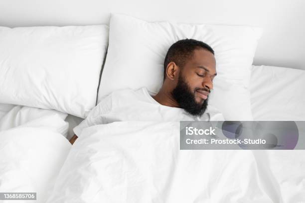 Calm Millennial Black Bearded Male Sleeping On Bed Soft Pillow With White Duvet In Bedroom Enjoy Rest Stock Photo - Download Image Now