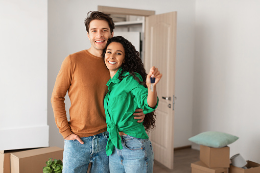 House Ownership. Portrait Of Happy Young Couple Holding Showing Key Standing In New Flat, Cheerful Guy Embracing Lady Posing After Moving In Own Apartment. Insurance, Real Estate, Mortgage Concept