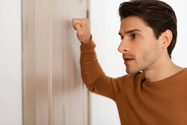 Closeup portrait of young handsome man knocking on the wooden closed door, male visitor standing in front entrance, guest wants to come in, free copy space, selective focus on face