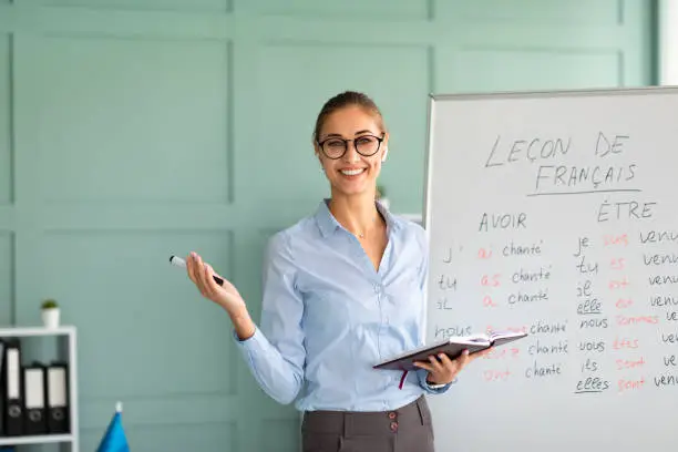 Photo of Friendly happy teacher recommending foreign languages school, giving French lesson, standing near whiteboard and smiling