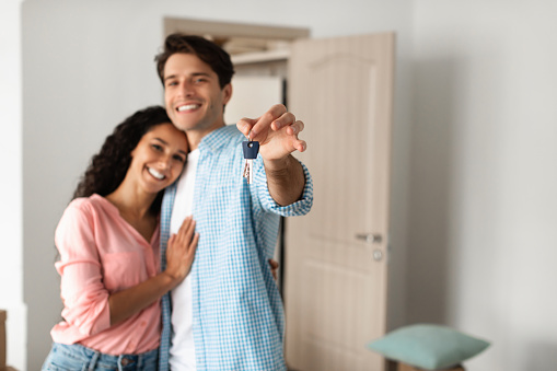 Happy millennial family holding and showing key standing in their new apartment flat. Cheerful guy and young lady moving to own house, selective focus on hand, blurred background, free copy space