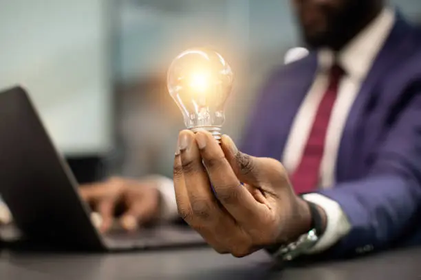 Photo of Black businessman holding illuminated light bulb, sitting in office