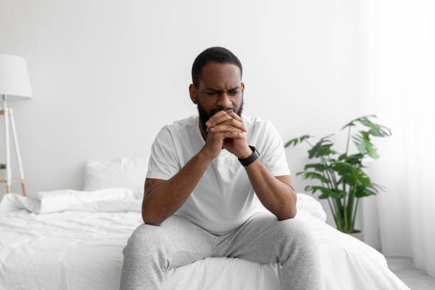 istock Depressed upset young black bearded male in white clothes sits on bed, think about solution difficulties 1368589638