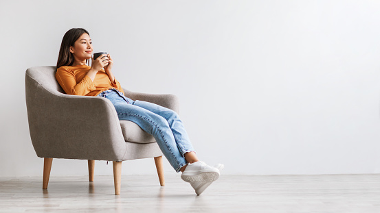Relaxed millennial Asian woman drinking coffee, having break, chilling in armchair against white studio wall, panorama with free space. Lovely young lady chilling and enjoying hot drink