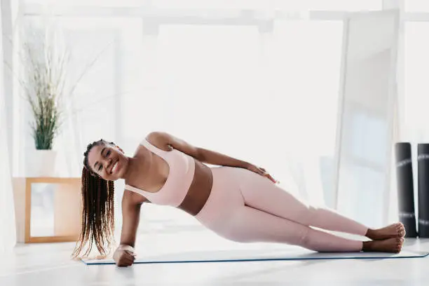 Photo of Beautiful young black woman standing in side plank on yoga mat at home, free space