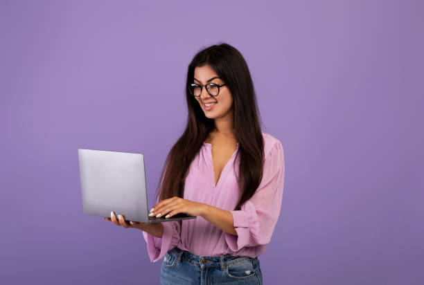 happy armenian lady using laptop computer and working remotely, posing over purple studio background - armenian ethnicity imagens e fotografias de stock