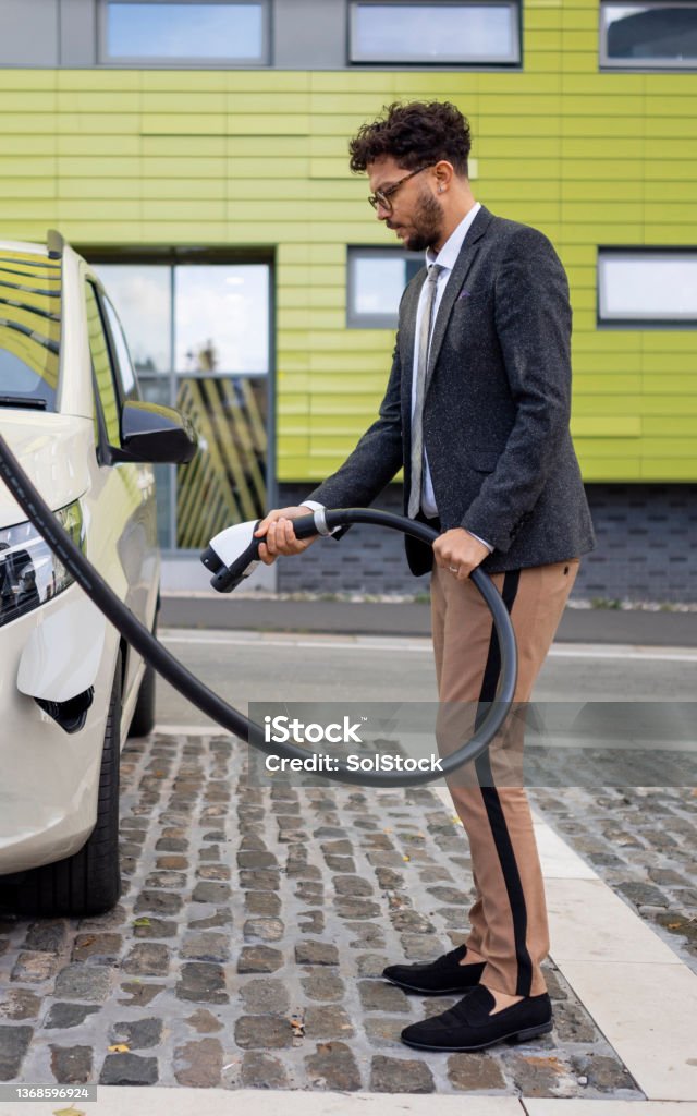 Charging My New Car A wide angle full length side view of a man in business dress putting his electric minivan on charge at a public charging point in Newcastle upon Tyne in the North East of England. Charging Stock Photo