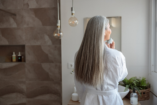 Back view of beautiful mature woman with long hair in white bathrobe standing in front of mirror and massaging face skin using massager