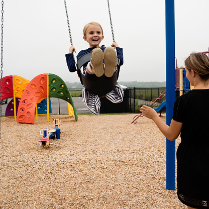 Cute blonde little girl playing in swings in public playground on a chilly and grey summer day. Young adult babysitter watching. Little girl is 4 year’s old and she is wearing a dark blue and white dress with stripes. Square full length outdoors shot with copy space.