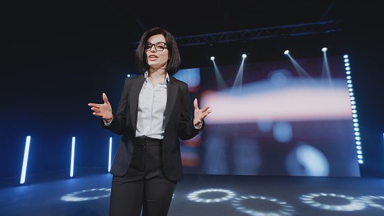 Female successful speaker on stage explaining to the audience investments and exchanges in front of an LED screen with 3D bitcoin in a room with spotlights, during a cryptocurrency training
