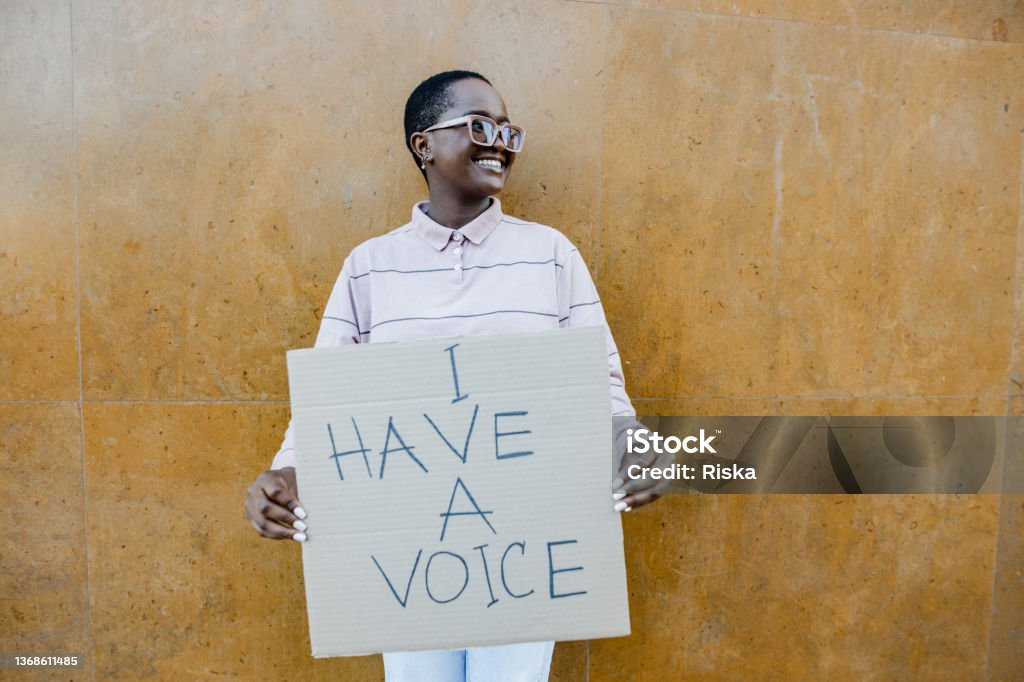 Portrait of woman holding anti racism sign Young woman in front of the wall, standing and holding a sign "I have a voice" Protest Stock Photo