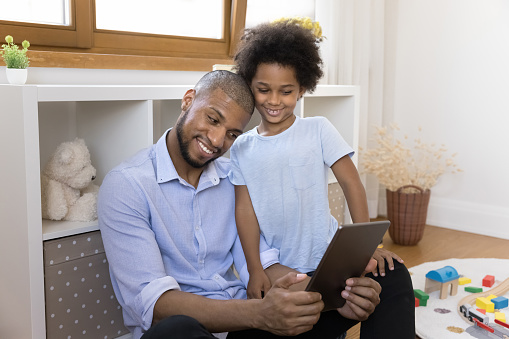Happy African American dad and little son kid watching funny media content on tablet together, making video call. Loving father teaching boy to use online learning app, digital gadget for studying