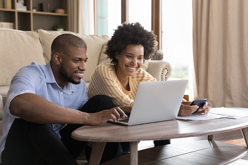 Happy young Black couple using laptop, looking at screen, smiling, laughing, getting good news. Husband and wife using online banking app on computer, paying bills, insurance mortgage fees on internet