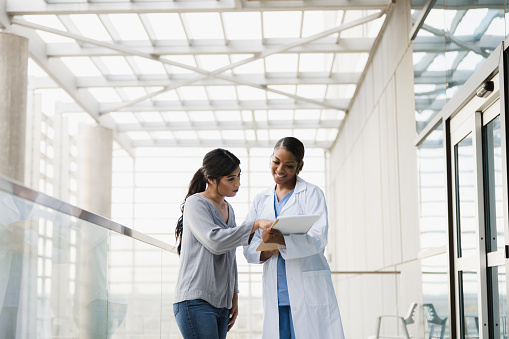 Standing in the walkway outside the patient rooms at the hospital, the mid adult female doctor gives the young adult female family member good news.