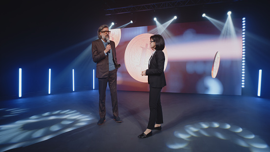 A female presenter in a suit interviewing a cryptocurrency coach on stage in front of an LED screen with a 3D object in a room, with spotlights during investment event