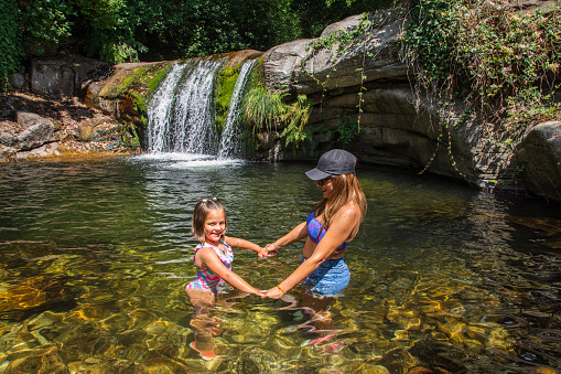 Mother And Daughter Bathing In A River With A Beautiful Waterfall In Nature. Young Woman With A Small Girl Enjoying In A Natural Pool Of A River With Transparent Water In A Town Of Cáceres, Spain.