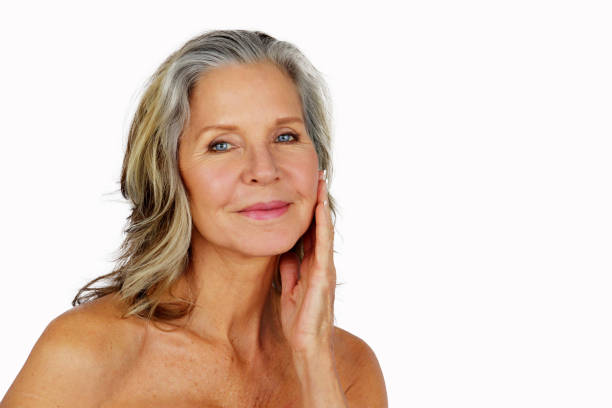 Beautiful older woman touching left side of face with open hand and smiling.  She has shoulder length greying hair