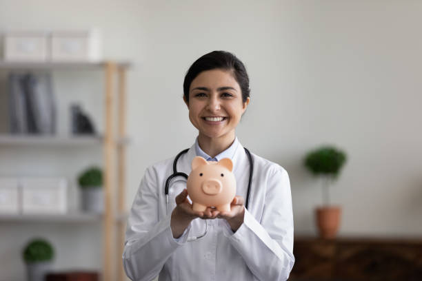 Head shot portrait smiling Indian female doctor holding piggy bank Head shot portrait smiling Indian female doctor physician in white uniform with stethoscope holding pink piggy bank, looking at camera, medical insurance or charity concept, healthcare and medicine operating budget stock pictures, royalty-free photos & images