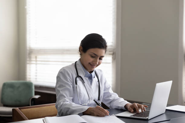 istock Smiling Indian woman doctor taking notes, using laptop in office 1368636270